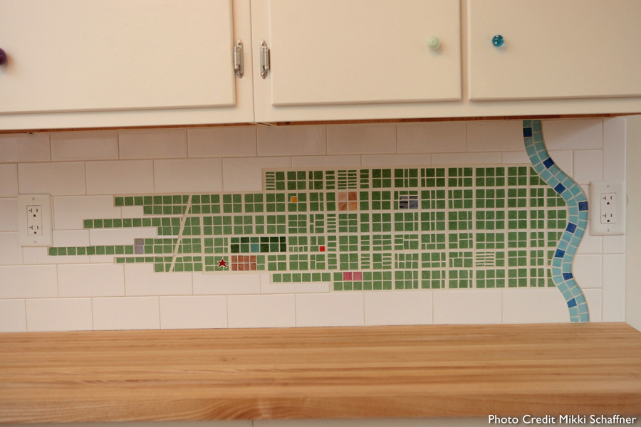Kitchen backsplash featuring a green glass tile mosaic map of historic 1842 Cincinnati, with a wavy iridescent blue Ohio River along the right edge, under white cabinets with mismatched colorful glass knobs