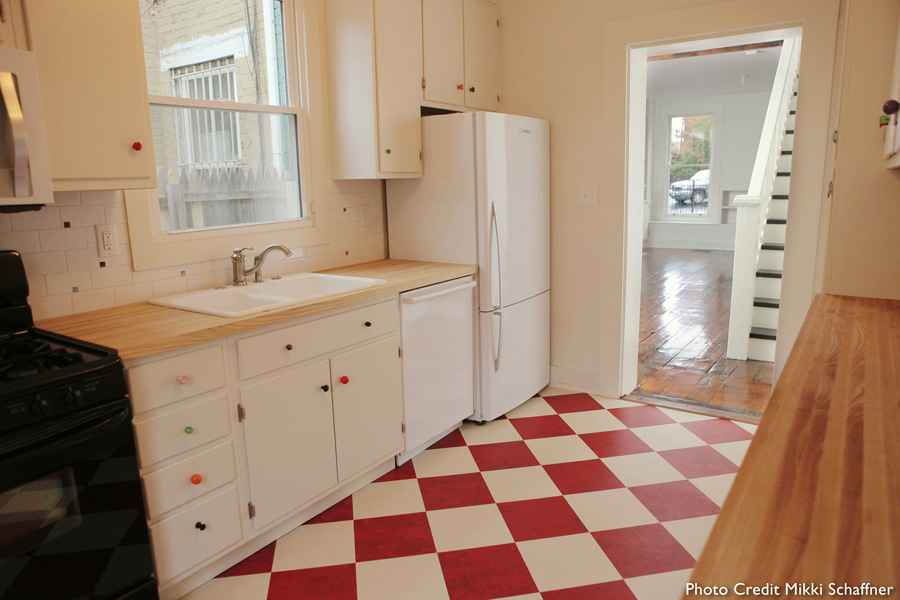 Galley kitchen with red-and-white checkered tile floor, butcher block counter, white cabinets with mismatched colorful glass knobs, gas stove, and side-by-side refrigerator, stairs visible through doorway