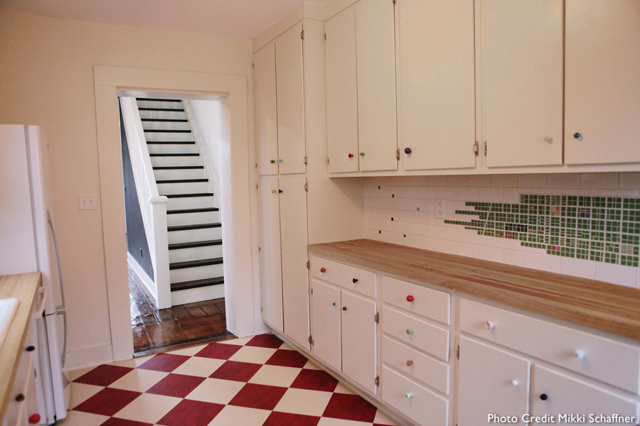 Long view of a galley kitchen with the green-tile 1842 Cincinnati map backsplash, butcher block counter, white cabinets with multicolor glass knobs, and red-and-white checkered tile floor