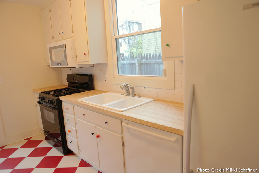 Kitchen sink and stove area — white drop-in sink, butcher block counter, white cabinets with colorful glass knobs, gas range, window over sink, red-and-white checkered tile floor