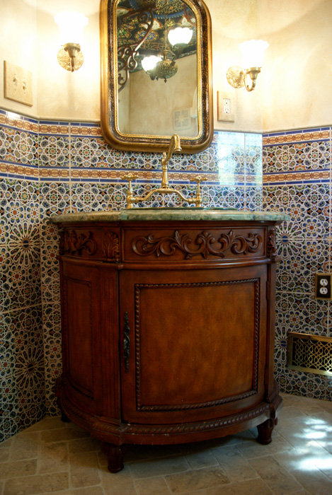 Vanity with carved wood cabinet, green marble countertop, gilded brass faucet, ornate gilded mirror, wall sconces, and walls clad in blue, yellow, and red Moroccan geometric pattern tile
