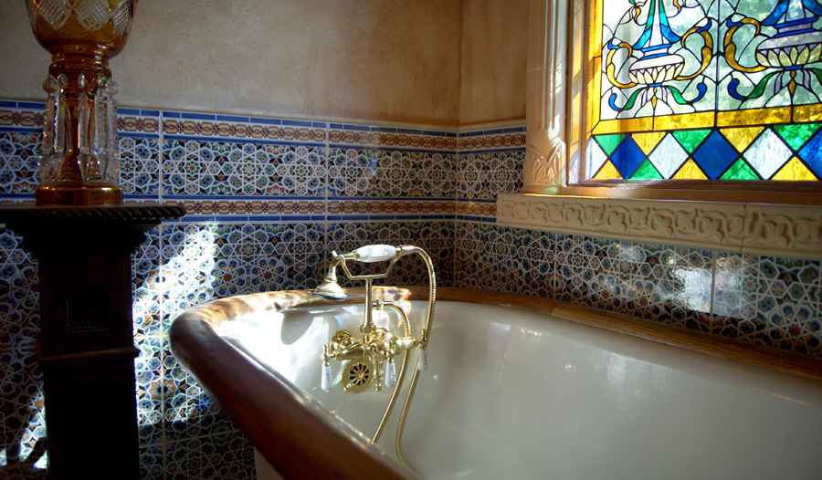 Claw-foot tub with gilded brass faucet, Moroccan pattern tile walls, and a stained glass window featuring blue, green, and amber Art Nouveau floral motifs