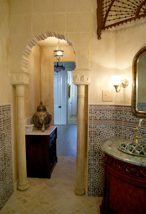 Moorish arched doorway with carved white capitals and columns, hanging lantern, hand-painted plaster walls, intricate carved wood screen above, and Moroccan tile wainscot