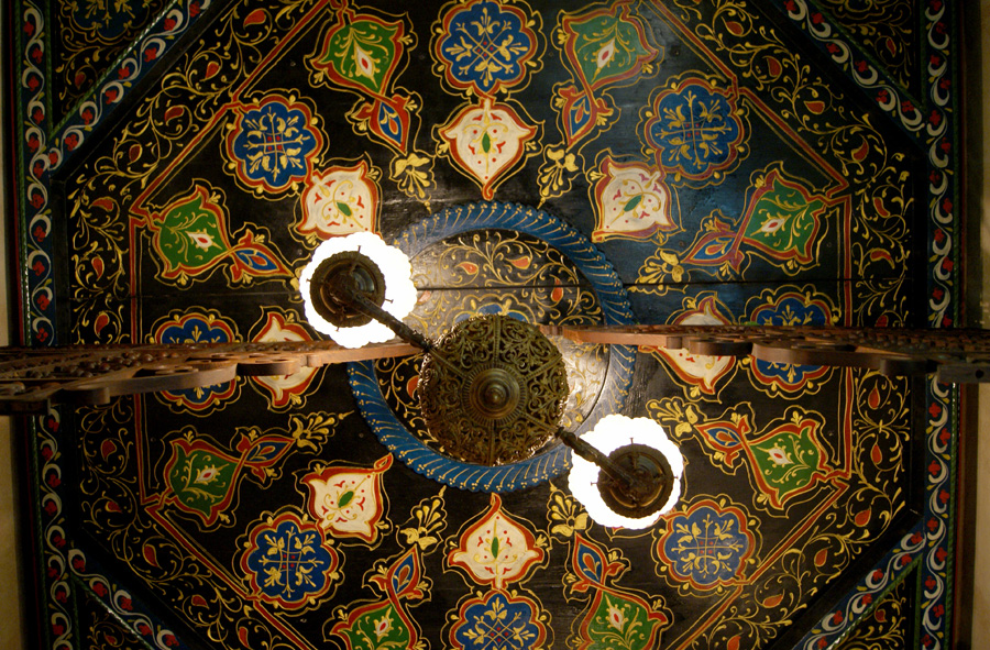 Hand-painted ceiling viewed from below — dark blue ground with intricate gold, red, and green decorative panels in geometric and floral patterns, and a central chandelier with twisted glass arms