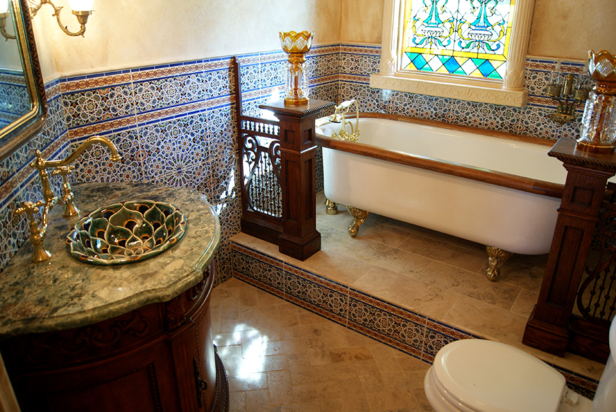 Moorish-inspired bathroom with white claw-foot tub on gold feet, carved wood pedestal holding a crystal candelabra, vanity with green marble counter and decorative copper bowl sink, Moroccan tile, and travertine floor