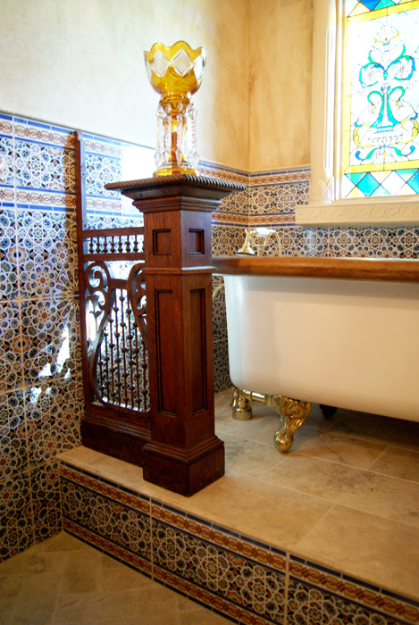 Detail of carved wood pedestal with iron filigree screen panel and a crystal candelabra above, claw-foot tub with gold feet, Moroccan pattern tile wainscot, and a stained glass window beyond