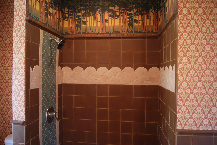 Shower wall with a Bradbury-style Pine Stands forest frieze along the top, brown rectangular tile with a pink scallop-tile band, and a sage green herringbone column with oil-rubbed bronze shower head