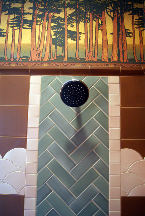 Looking up at a shower wall — Pine forest frieze above a brown tile field, sage green herringbone column with oil-rubbed bronze shower head, and pink scalloped fish-scale tile trim
