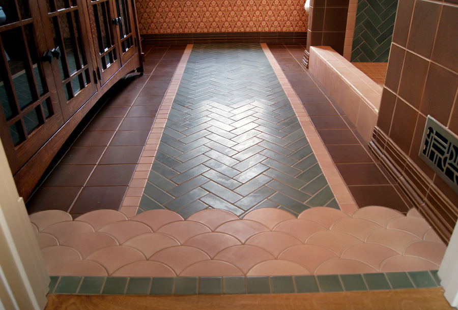 Bathroom floor with sage green herringbone runner bordered by pink scalloped fish-scale tile at the threshold, brown tile field, Mission-style oak vanity, dark wood paneling, and Arts & Crafts wallpaper