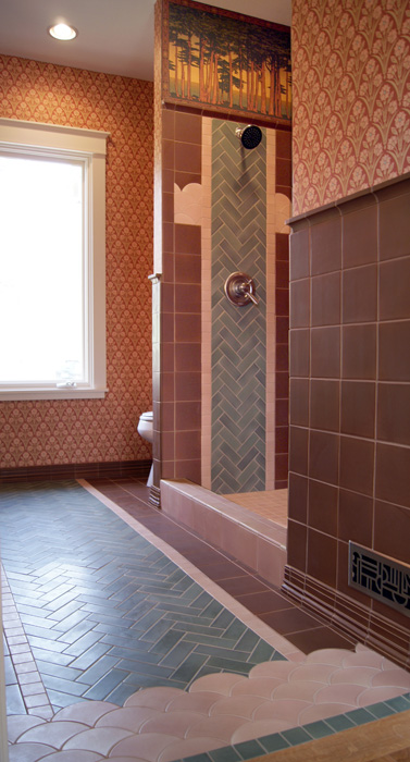 Side view of an Arts & Crafts shower stall — brown porcelain tile walls, sage green herringbone center column with shower head, pink scallop-tile band trim, and a brown herringbone floor outside the shower