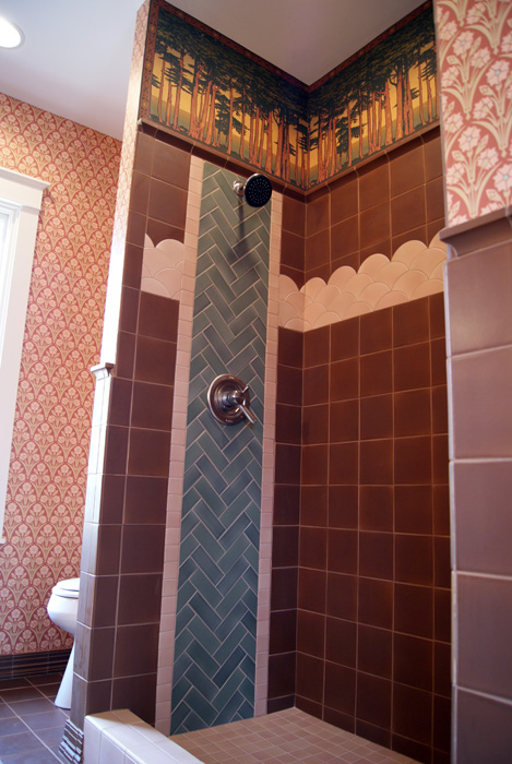 Arts & Crafts shower stall with a Pine Stands forest frieze along the top, sage green herringbone column with shower fixtures, pink scallop-tile band trim, and a brown porcelain tile field