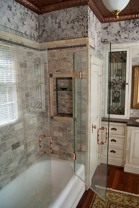 Shower stall with gray tumbled travertine brick wall tile, glass shower doors with polished copper grip handles, a recessed niche with copper trim and glass shelf, and an embossed tin ceiling above