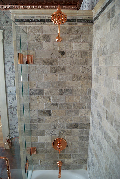 Shower interior detail — gray tumbled travertine wall tile, polished copper shower head with rope-twisted arm, copper accents, and a decorative leafy band trim