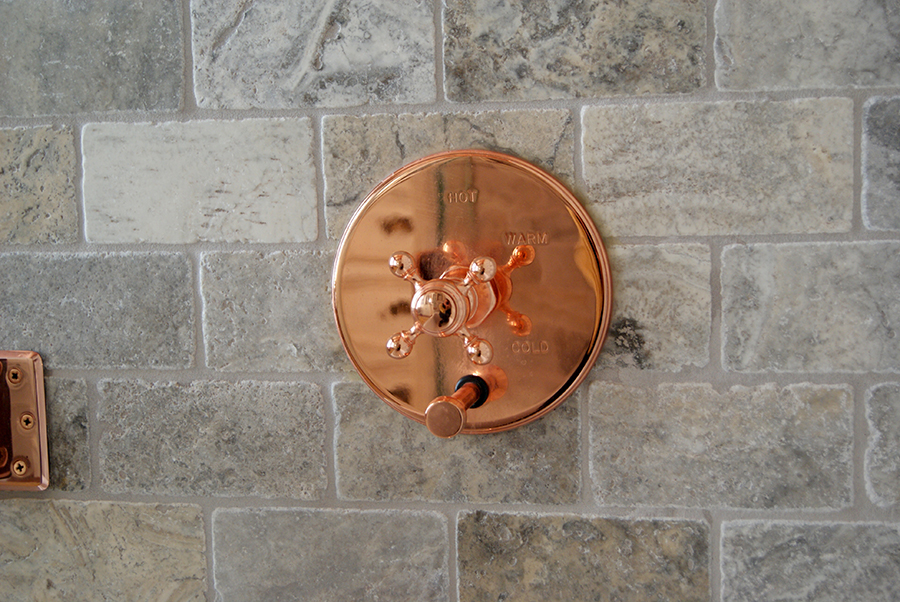 Close-up of a polished copper shower control valve engraved with hot, warm, and cold dial markings, set against gray tumbled stone brick wall tile