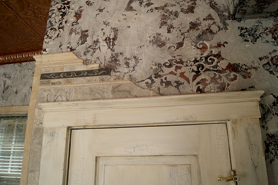 "Falling stones" tile detail — gray stone tile cascading from a cornice molding above a distressed cream-painted door frame, with a decorative leafy band tile, set against damask wallpaper