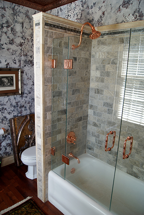 Side view of a tub-shower — distressed cream column with stacked stone tile detail, glass shower wall with copper bar pulls, polished copper shower head, gray stone tile walls, damask wallpaper, and an antique carved chair