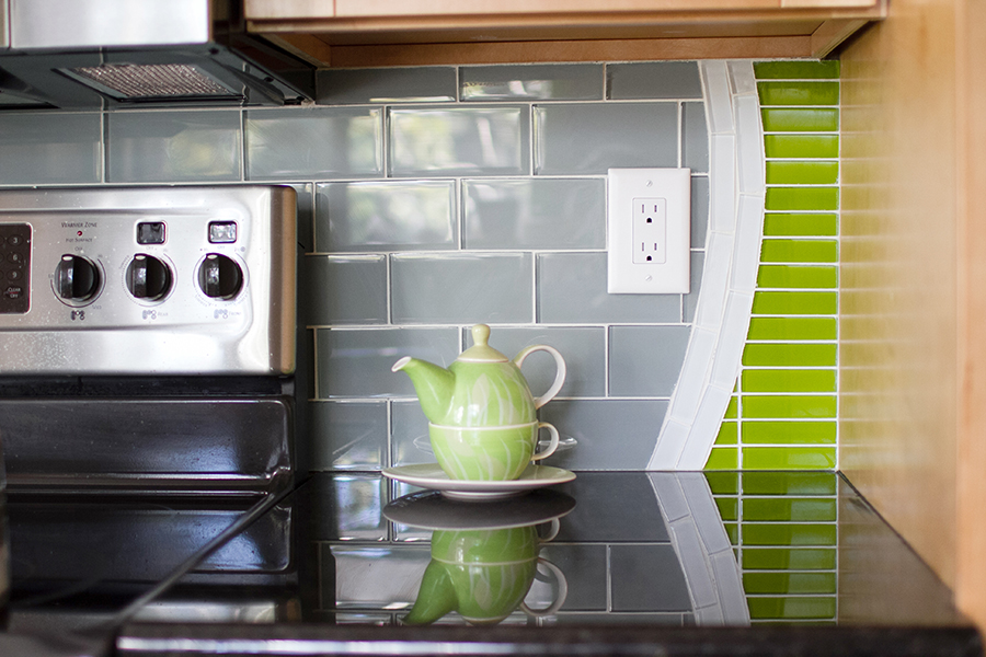 Kitchen stove area with stainless gas range, gray glass subway tile backsplash, lime green linear glass mosaic curving down the right side, and a lime green teapot on black granite counter