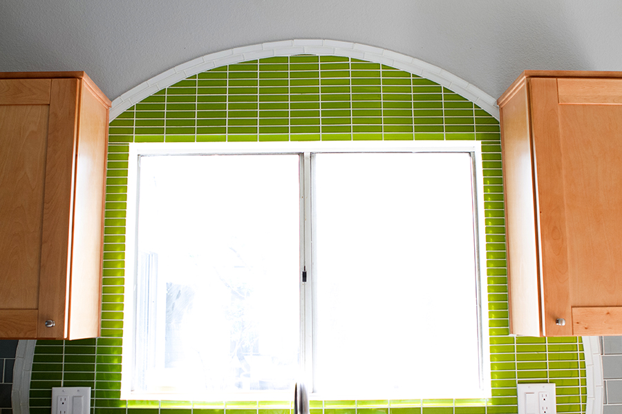 Kitchen window over sink with an arched lime green linear glass mosaic surround tracing the arch, maple shaker cabinets on either side, and gray painted walls