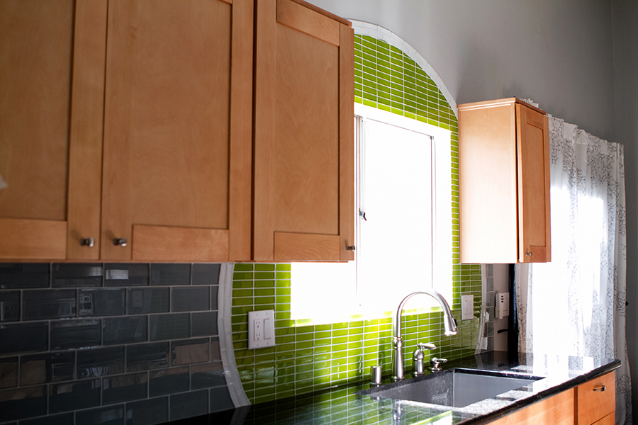 Kitchen sink view with an arched lime green linear glass mosaic feature over the window, gray glass subway tile backsplash, stainless undermount sink, maple cabinets, and black granite counters