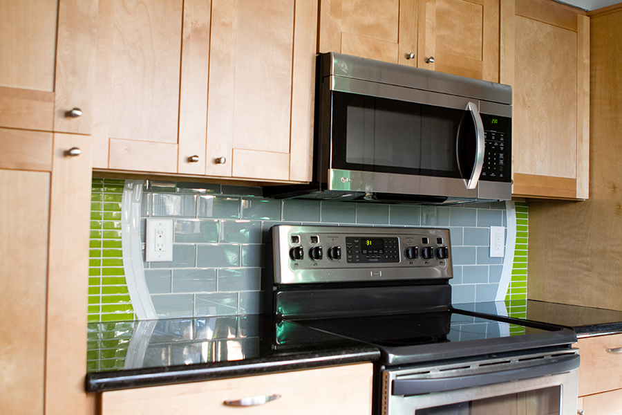 Kitchen stove and microwave area — stainless range hood/microwave, maple cabinets, gray glass subway tile field with lime green linear mosaic curving along the edges, and black granite counter
