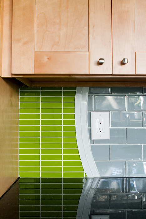 Detail of a curved tile transition between lime green linear glass mosaic and gray glass subway tile, beneath maple shaker cabinets