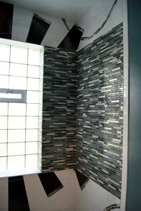 Shower interior with glass block window meeting a wall of vertical blue, teal, and silver linear glass mosaic, with dark rust tile rays and curved mosaic arc above
