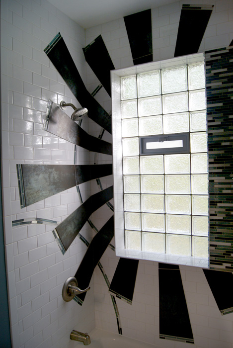 Tub-shower with central glass block window and dark rust tile rays radiating in a sunburst pattern across white subway tile, with blue glass mosaic accents
