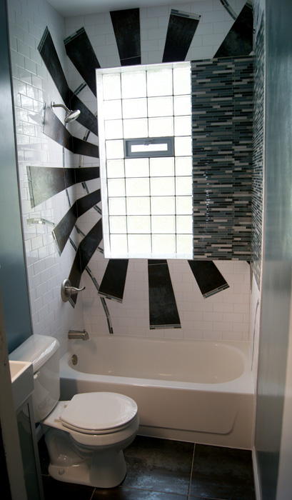 Bathroom with tub-shower beneath a glass block window, dark rust tile sunburst pattern radiating across white subway tile, vertical blue glass mosaic accent strip on the right wall