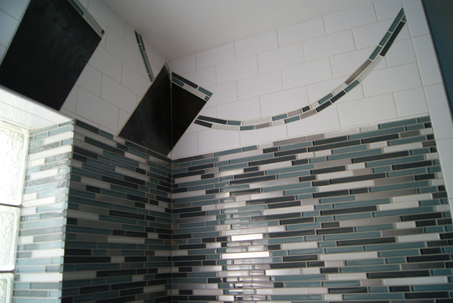 Detail of a shower wall — white subway tile, dark rust tile rays, curved arc of blue and silver glass linear mosaic, with a vertical multicolor glass mosaic field on the side wall