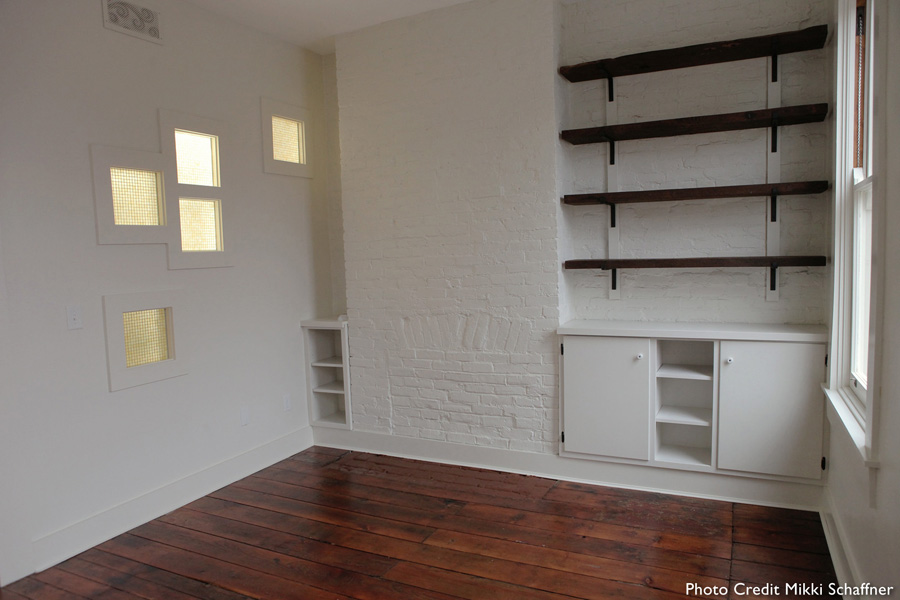 Open room in a tiny row house renovation with white walls, white-framed inset windows, white-painted brick column with dark wood floating shelves, and refinished dark wood plank floors