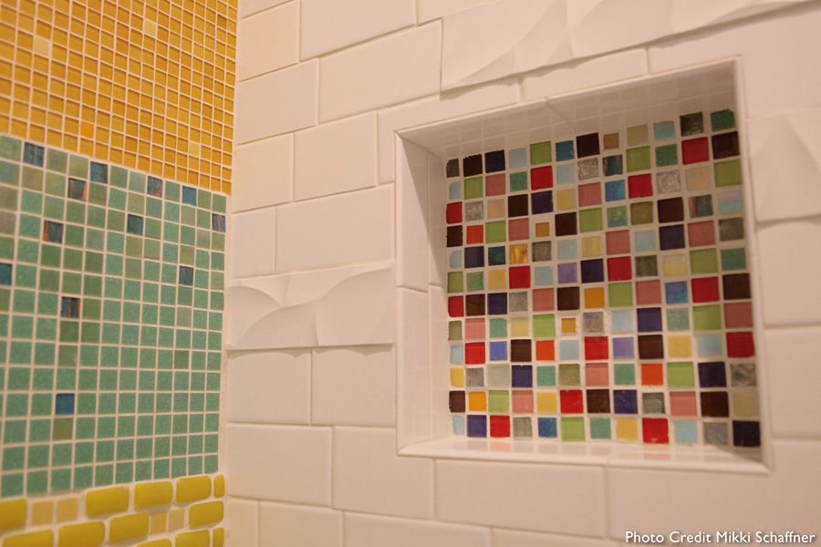 Bathroom wall detail — colorful patchwork mosaic meeting white subway tile with sculpted wave-pattern accent tiles, a small recessed niche lined with multicolor mosaic, and a travertine carved accent square