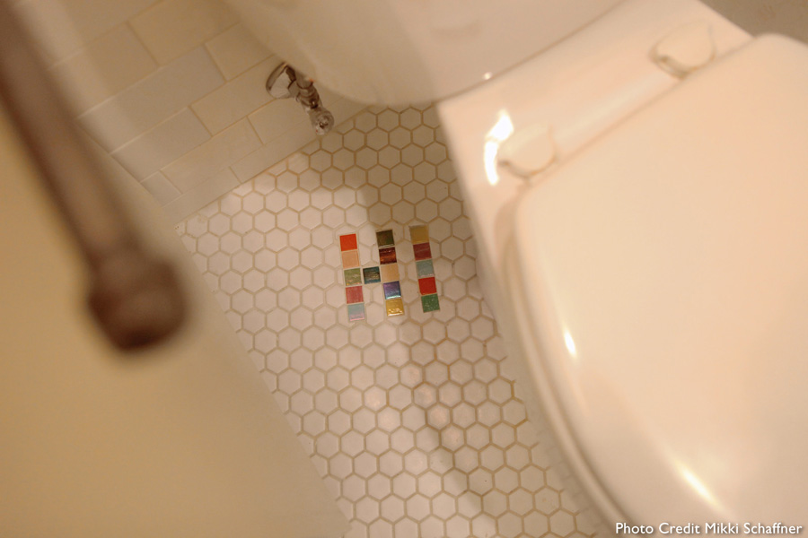 White hexagonal floor tile with a small "H"-shape laid out in colored hex tile accents, base of toilet visible