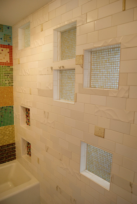 Bathroom wall with white subway tile, sculpted wave-pattern accent tiles, multiple recessed niches lined with multicolor mosaic, and the patchwork quilt mosaic visible on the side wall