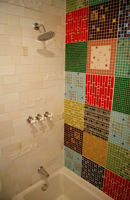 Shower interior with chrome fixtures — white subway tile with sculpted wave detail on the left wall, the multicolor patchwork quilt mosaic on the right wall