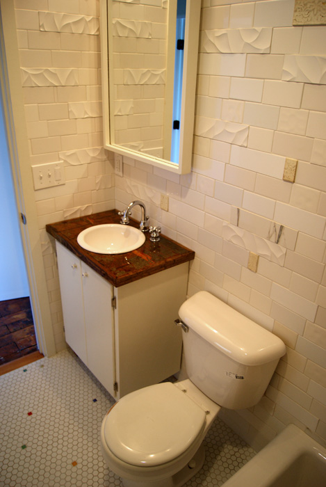 Bathroom vanity with white drop-in sink, dark copper-patina countertop, mirror, white subway tile walls with sculpted wave-and-ribbon detail tiles, and white hex floor with scattered colored accent tiles