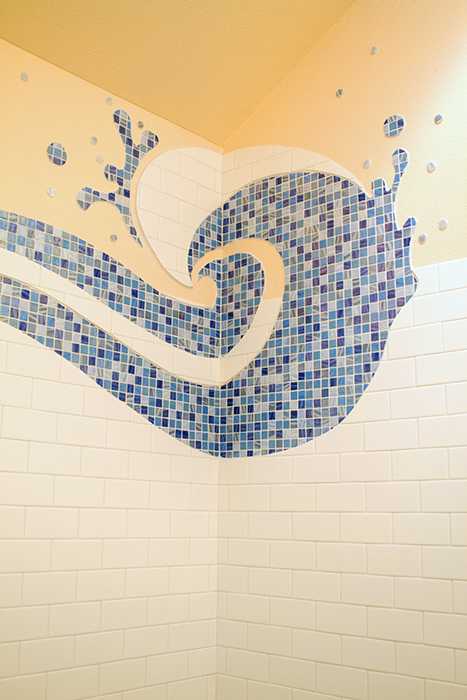Corner of a shower with a curling blue glass mosaic wave and splash droplets sprayed above, white subway tile field below, pale yellow wall