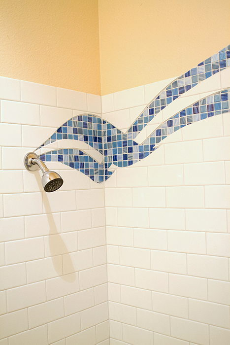 Chrome shower head with the tail of a blue glass mosaic wave flowing into it, two parallel curving mosaic ribbons above, white subway tile