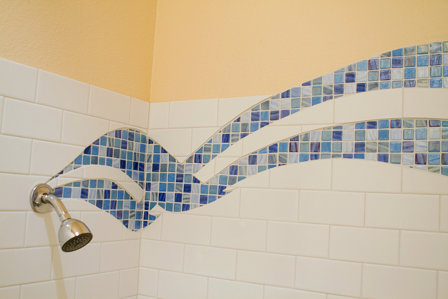 Detail of multiple parallel curving blue glass mosaic ribbons in varied blue tones flowing into a chrome shower head, white subway tile field