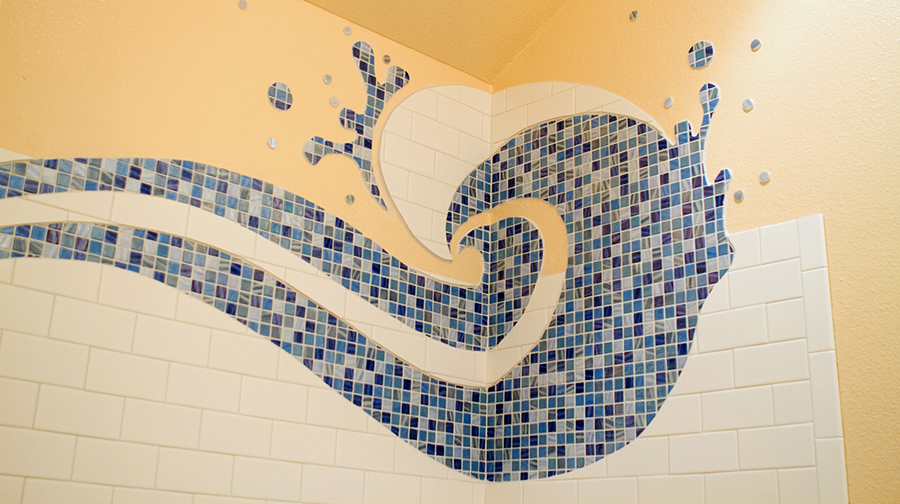 Detail of a cresting blue glass mosaic wave curl with droplets and spray pattern, pale yellow wall above white subway tile