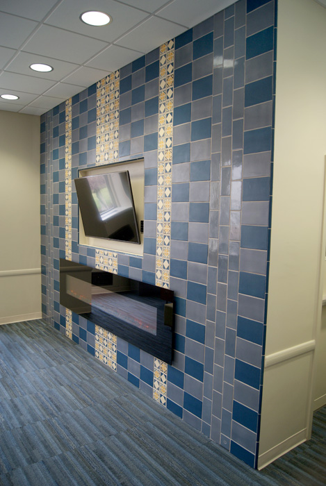 Angled view of a Rookwood tile media wall — checker pattern of blue and gray-blue field tile with decorative cream and gold pattern inlay columns, TV and fireplace