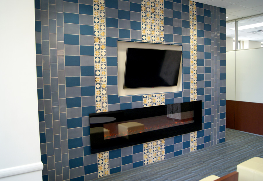 Office lounge media wall — Rookwood tile in alternating blue and gray field with decorative quatrefoil inlay tile columns, framing a mounted TV and linear gas fireplace