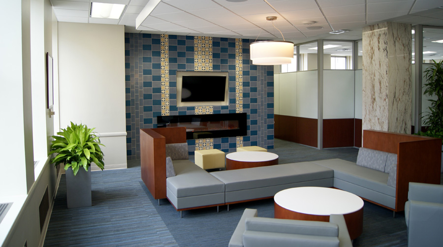 Office lounge with a Rookwood tile media wall installation, modular gray sofa seating, drum pendant light, floor plant, and a marble pillar in the foreground