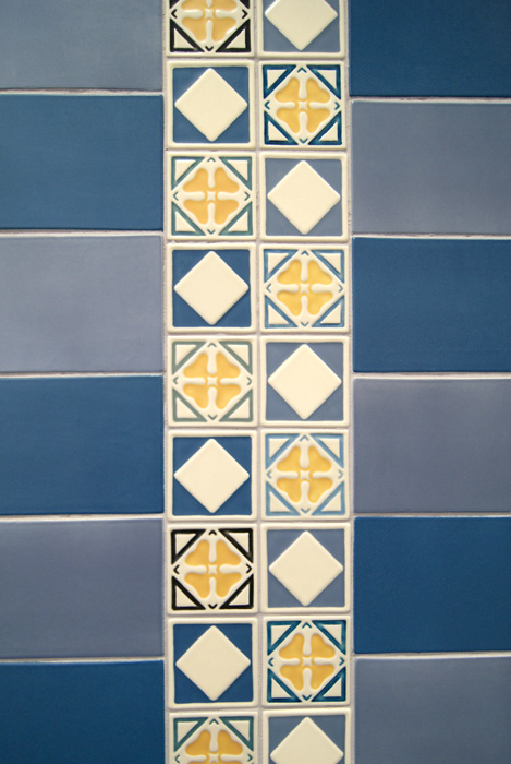 Close-up of decorative Rookwood inlay tiles — cream and blue squares with gold quatrefoil pattern motifs and triangular corner accents, set between blue and gray-blue field tile