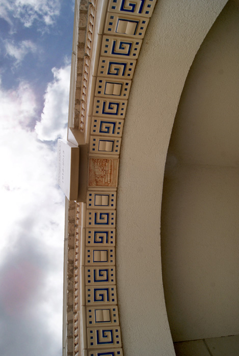 Looking up at an arched library entryway with a Rookwood tile band of Greek key pattern in cream and dark blue, dentil molding above, cloudy sky beyond