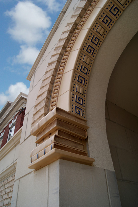 Detail of an arched library entry — Rookwood tile band with cream and blue Greek key pattern curving along the arch, dentil molding above, and a stacked-book-style decorative corbel below the springer