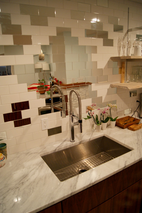 Kitchen sink area with white subway tile backsplash dotted with small mahogany and silver mirror accent tiles in a checker-like pattern, stainless undermount sink, marble counter, chrome gooseneck faucet, and orchids