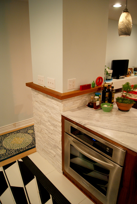 Kitchen corner with stacked white stone veneer column meeting marble counter, integrated stainless wall oven, and a glass pendant light overhead