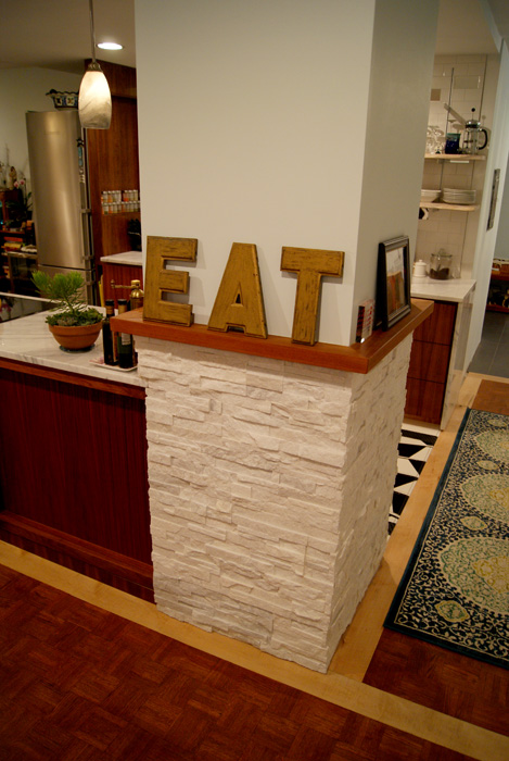 Kitchen island wrapped in stacked white stone veneer with a wood top displaying wooden "EAT" letters, oriental rug on hardwood floor
