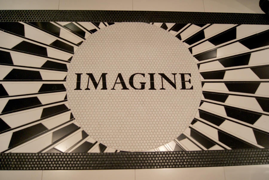 Detail of the "IMAGINE" floor mosaic — black-and-white penny tile circular medallion spelling IMAGINE in serif type, framed by radiating starburst rays of black ceramic tile