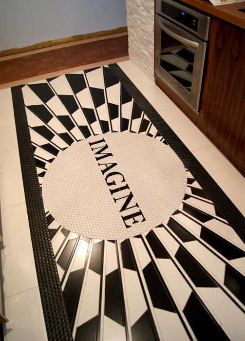Angled view of the "IMAGINE" penny-tile floor mosaic between kitchen cabinets and a stacked stone column with integrated wall oven, starburst pattern radiating from the central medallion